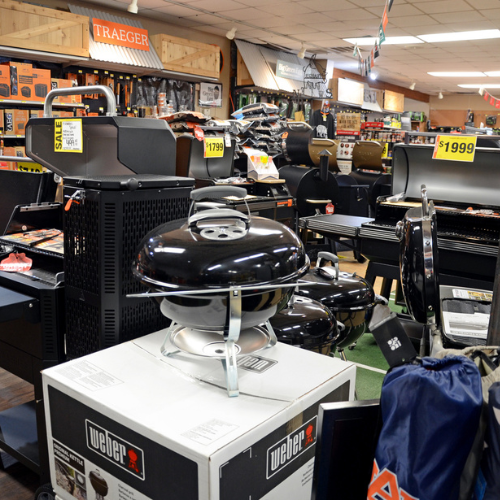 A store interior with various barbecue grills on display. Prominent brands like Weber and Traeger are featured. Sale signs show prices, creating a busy retail atmosphere.