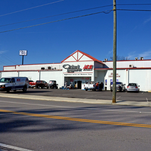 Large hardware store with a red and white facade, several cars, and a van parked in front. Clear blue sky and power lines above. Busy and welcoming.