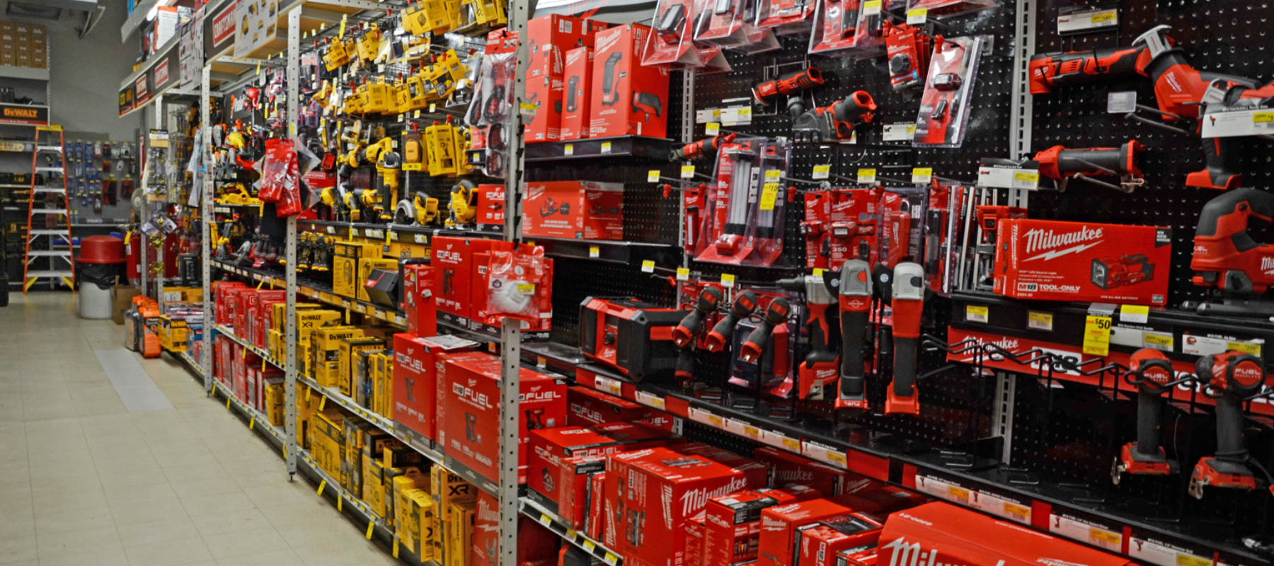 Hardware store aisle with shelves stocked with various power tools in red and orange packaging. Brightly lit, orderly display with a range of brands.